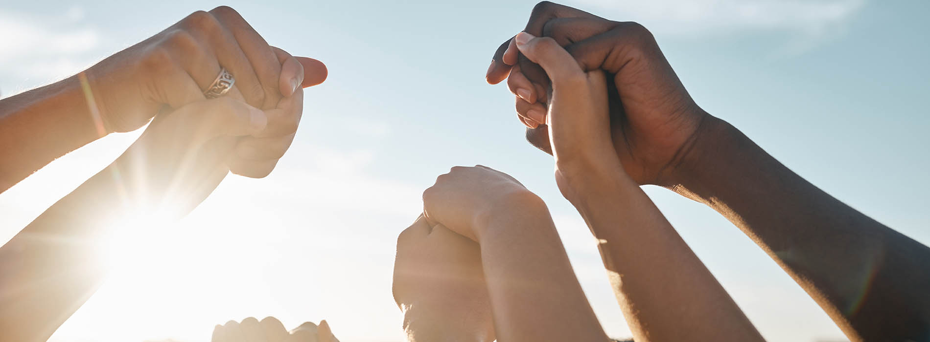The image shows multiple hands reaching upwards towards the sky with fingers interlocked, creating a sense of unity and connection, against a bright background that suggests an outdoor setting.
