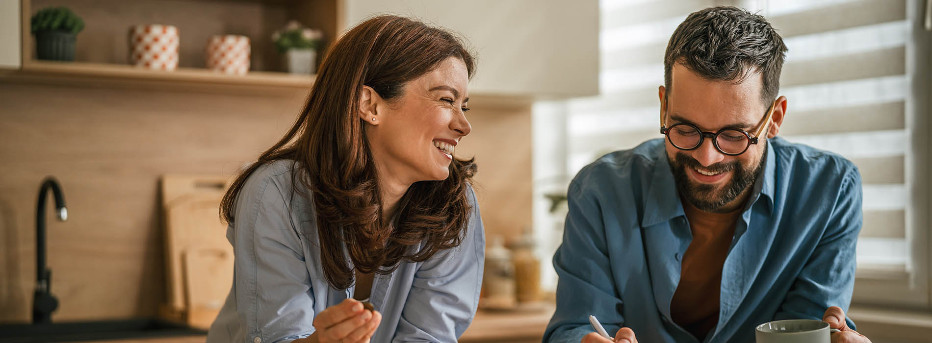 A man and woman are shown in a kitchen setting, sitting at a table with a laptop between them, both smiling and looking towards each other.