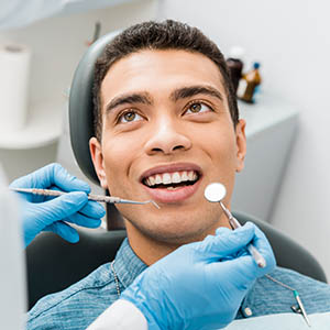 The image depicts a man sitting in a dental chair with his mouth open, receiving dental treatment from a professional who is cleaning his teeth with a toothbrush.