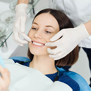 The image shows a dental hygienist performing a teeth cleaning procedure on a patient who is seated in a dental chair, with both of them wearing medical gloves and protective eyewear.