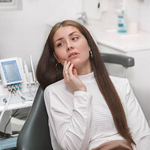 The image shows a woman with long hair sitting in a dental chair, looking down and to her right with a hand on her chin, while wearing a white top.