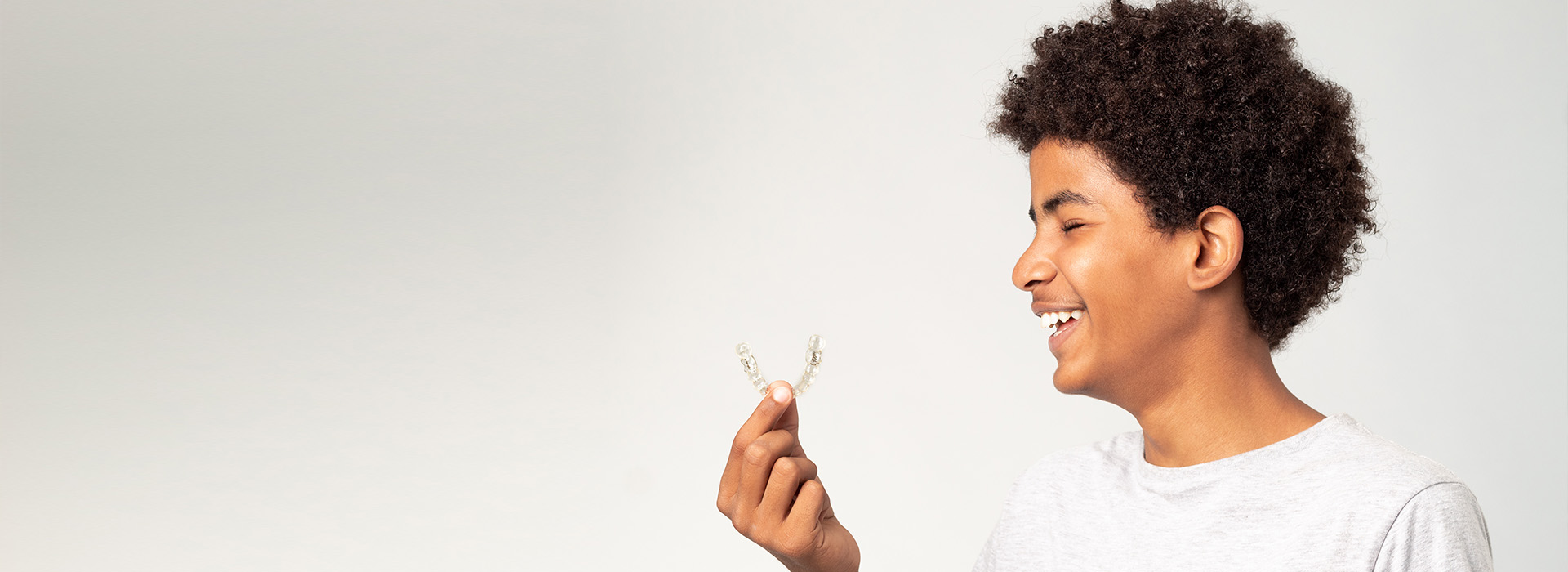 The image shows a person with curly hair holding a small object close to their face, smiling at the camera, against a plain background.
