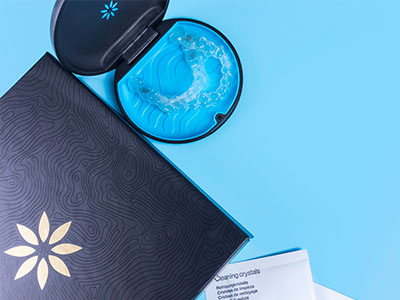 A close-up view of a blue box with a white design on top, placed next to an open blue container, possibly containing a product, on a blue surface with a black and gold patterned item in the background.