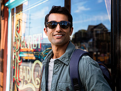 In the image, there s a man standing outside a shop, wearing sunglasses, with a backpack on his back, holding onto a railing, and smiling at the camera.