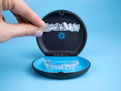 The image shows a person s hand holding a clear tray containing transparent dental aligners, with a close-up view of one aligner placed inside an open case displaying its shape.