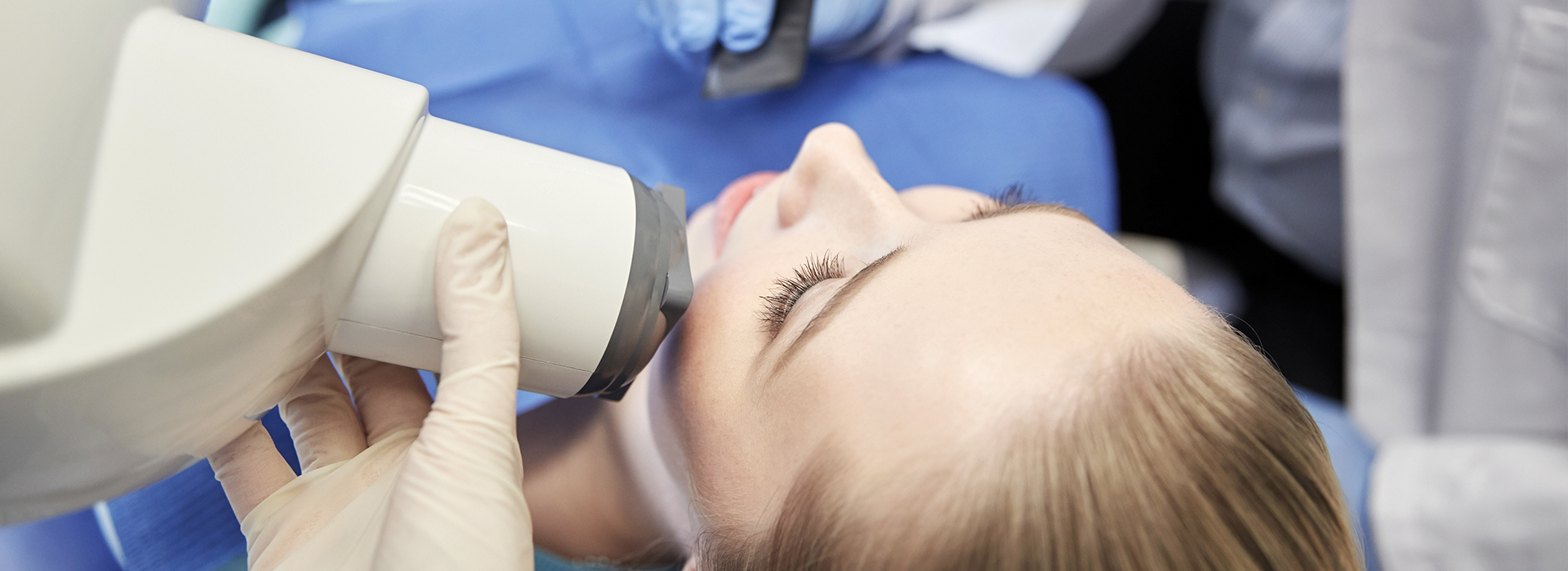 The image shows a person receiving dental treatment with a dental mirror and light held up close to their mouth by a professional, likely a dentist or dental hygienist, in an office setting.