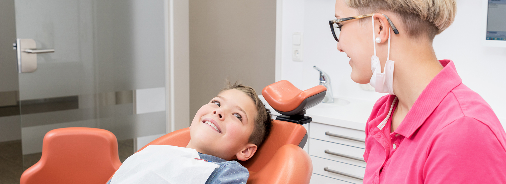 The image shows a dentist and patient in an office setting  the dentist is seated at a desk with a computer monitor, while the patient is sitting on a dental chair, smiling and looking towards the camera.