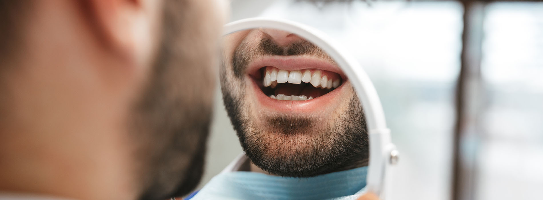 The image shows a close-up of a man with a medical mask covering his nose and mouth, smiling towards the camera, with a background that appears to be an interior space with natural light coming through a window.