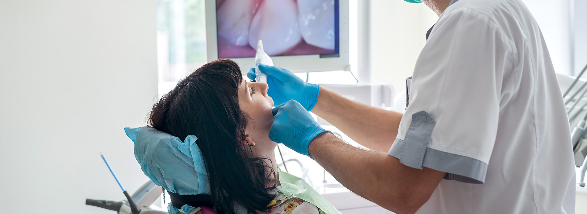 The image shows a dental professional performing an oral examination on a patient, with the dentist using a mirror and possibly other instruments to inspect the teeth and mouth.