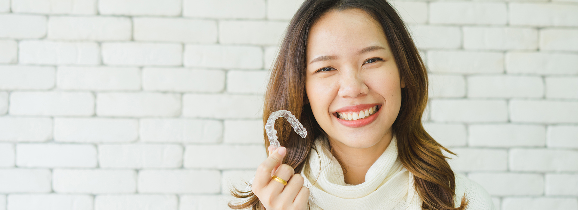 The image features a smiling woman holding a toothbrush in front of her face, with a brick wall in the background.
