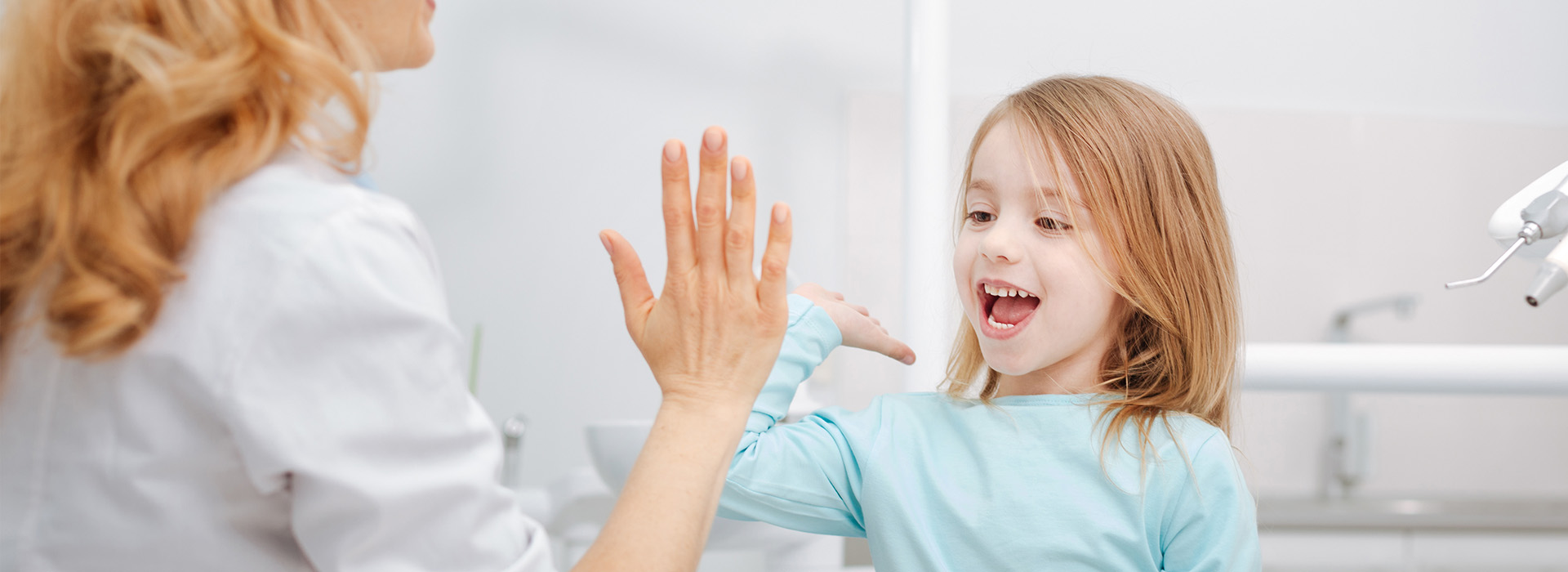 The image shows a young girl with blonde hair waving at the camera from behind a woman who appears to be washing her hands, both in an indoor setting with visible sinks and faucets.