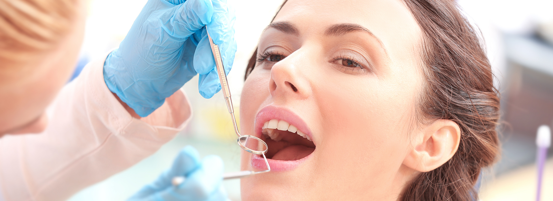 The image shows a woman receiving dental treatment, with a dentist using a drill on her teeth while she wears blue gloves and holds a mouth mirror.