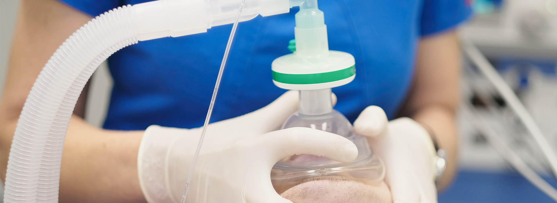 The image shows a medical professional wearing gloves and a mask, holding a clear plastic bottle with a green cap, which appears to contain a liquid or paste-like substance, possibly for medical use, in front of a blue background that might be part of medical equipment or a wall.
