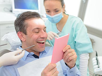 This is a composite image showing a man sitting in an oral surgeon s chair with a surgical mask on, holding a tablet with a smiling face on the screen, while looking at it with a surprised expression.