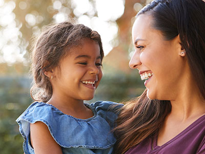 This is a photograph featuring an adult woman and a child smiling at each other  they appear to be outdoors during daylight, with the woman wearing a dark top and the child in a light-colored dress. The woman has long hair and is looking down at the child, who is standing in front of her.