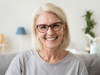 The image shows a smiling woman with short blonde hair, wearing glasses, sitting on a couch with a white wall in the background.