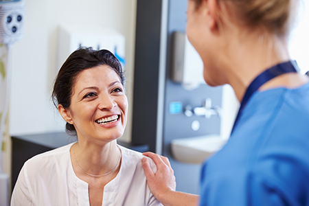 The image shows two individuals in a medical setting  one appears to be a woman wearing a white lab coat and stethoscope, smiling at another person who seems to be a patient.