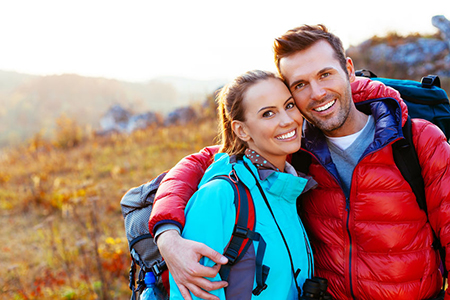 A man and woman smiling at each other while posing for a photo during an outdoor adventure, with the man wearing a backpack and both dressed in hiking gear.