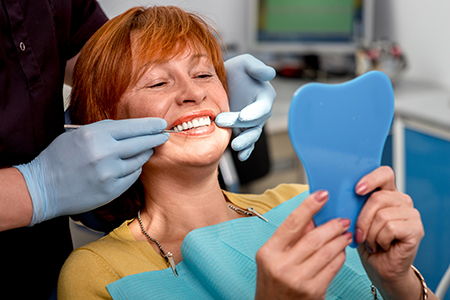 In the image, a woman is seated in a dental chair with a blue face mask on her face, holding up a toothbrush towards the camera while smiling. She appears to be engaged in a dental care activity or demonstration.
