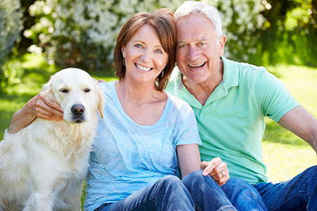 An elderly couple posing with their yellow labrador retriever on a sunny day.