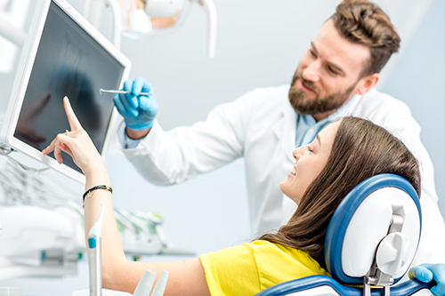 A woman wearing a blue headset, sitting in a dentist s chair, with a man in a white lab coat standing behind her, pointing at something on a computer screen.