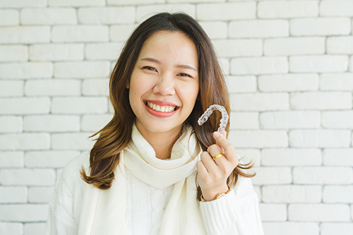 A smiling woman holding up a large candy cane with her right hand against a brick wall background.