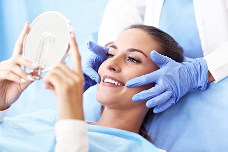 The image depicts a person seated on a dental chair with a mirror held up to their face, receiving attention from a dentist wearing gloves and a blue gown, who is performing a dental procedure.