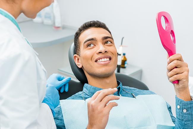 The image depicts a person sitting in a dental chair with a broad smile, wearing a blue shirt, and holding a pink object in their hand, while being attended to by a dental professional who appears to be adjusting something on the individual s mouth.