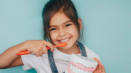 The image shows a young girl brushing her teeth with a toothbrush, holding a cup of toothpaste, and smiling at the camera while wearing a blue apron.