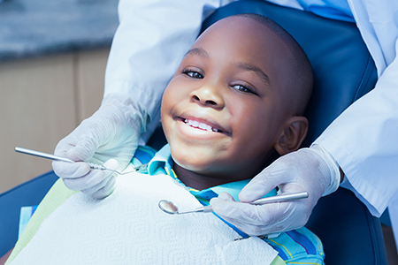 A young boy sitting in a dental chair with a smile on his face, receiving dental treatment from a dentist wearing white gloves and a mask.