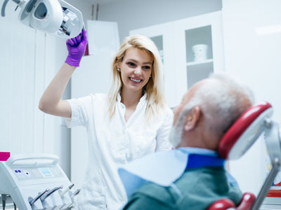 A woman with blonde hair wearing a white shirt and purple gloves is standing next to an older man wearing a hospital gown, who is seated in a dental chair with his eyes closed.