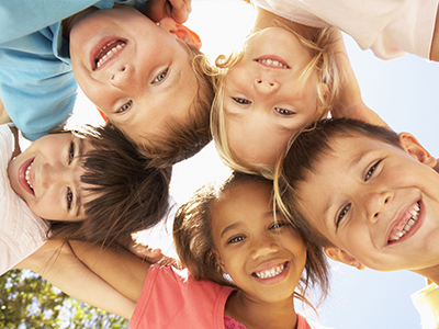 The image displays a group of children, all smiling broadly, posing for a photograph with their arms around each other, creating a circular formation.
